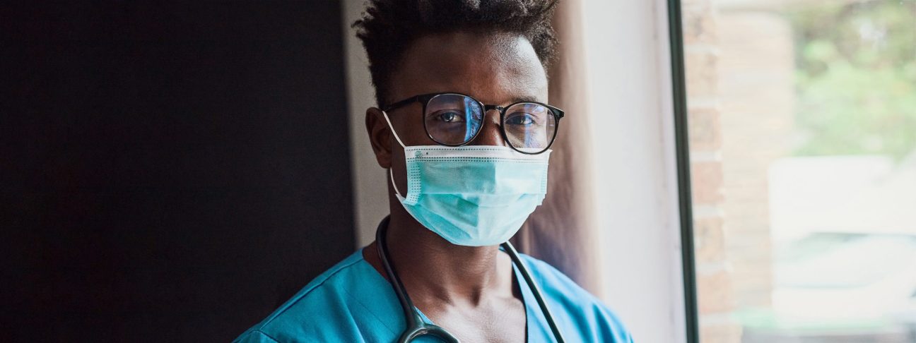 A male nurse in turquoise scrubs wearing round tortoiseshell glasses and a medical-grade face mask.